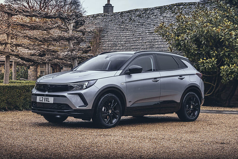 Silver modern SUV with black alloy wheels parked on a gravel driveway outside of an old stone cottage with a tiled roof and trimmed hedges, captured in a dynamic, side on view, under an overcast sky.