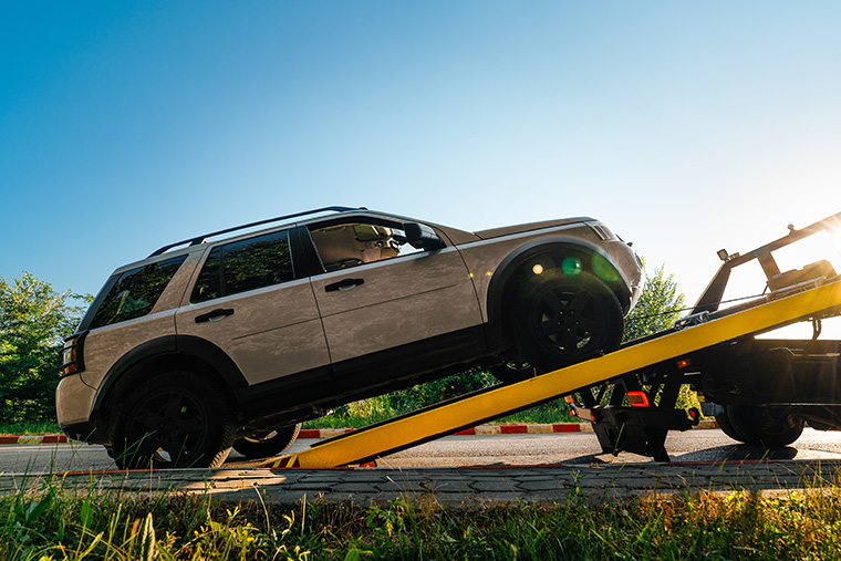 Beige SUV with black alloys being un-loaded off a yellow tow truck ramp on the side of a road, with greenery and a clear blue sky in the background.