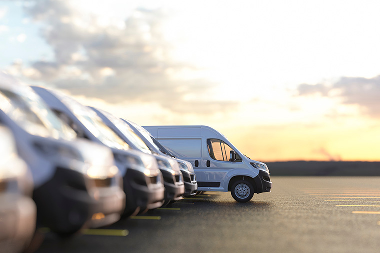 Row of white vans parked in a lot, with one van in the foreground clearly visible and in focus, while the others fade out of focus. The sky in the background shows a sunset with soft clouds.