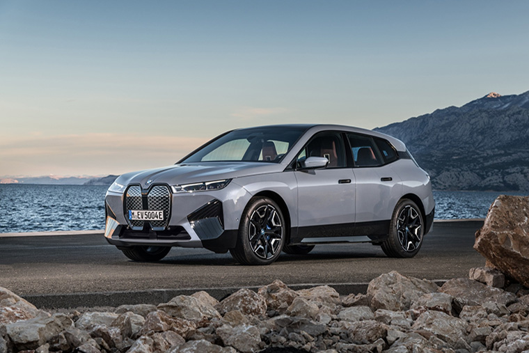 Silver BMW iX electric SUV parked on a road near the coast, with rocky terrain in the foreground and mountains in the background. The sky is clear, and the vehicle is captured in a dynamic, side-on view.