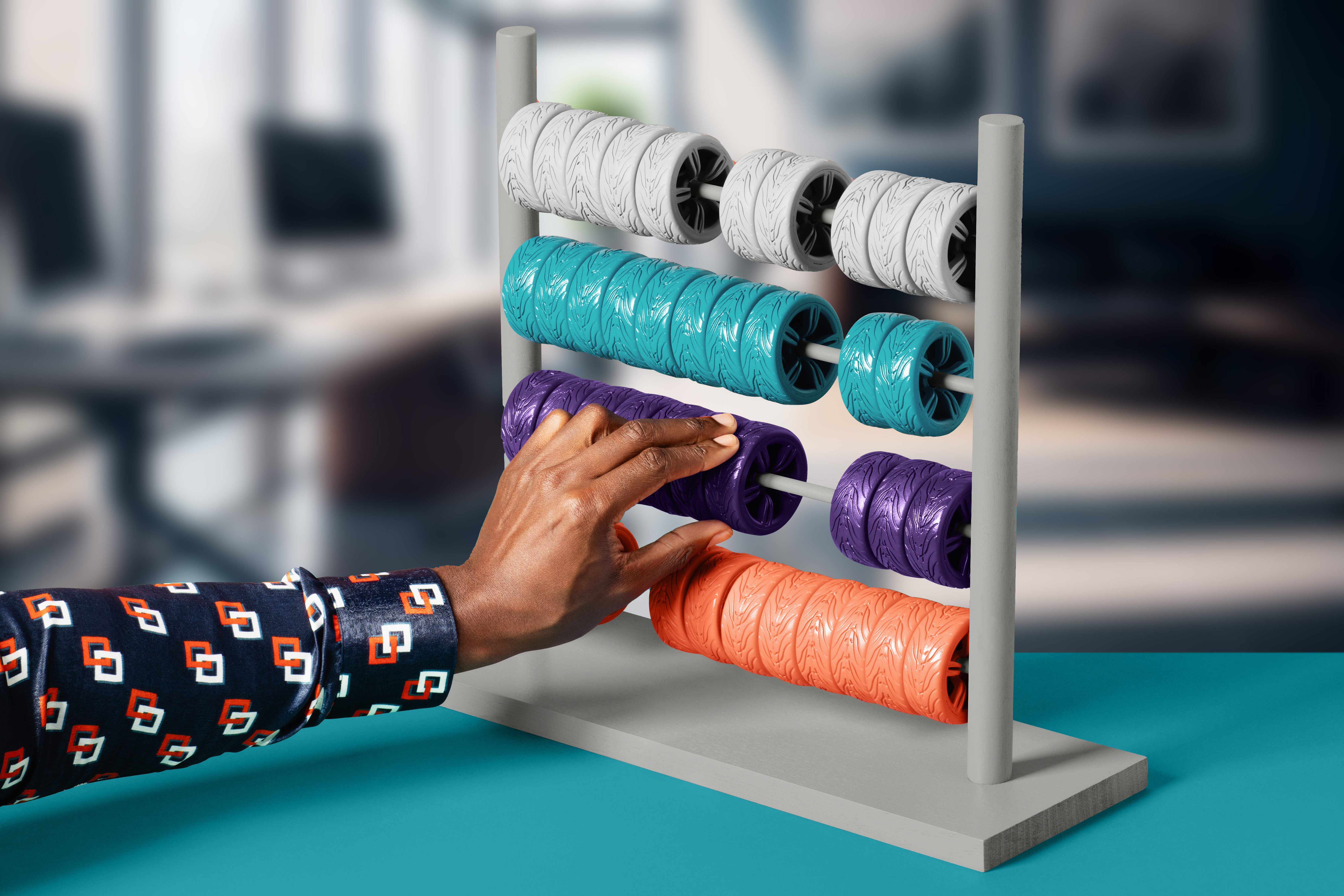 A hand adjusting a brightly coloured abacus with car wheels in white, teal, purple and orange, set against a blurred office background on a blue surface.