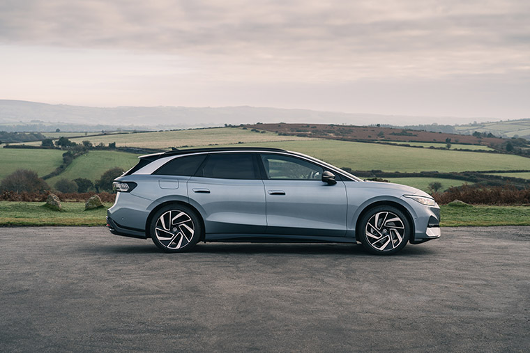 A side profile view of a silver Volkswagen electric car parked on a rural road with expansive green fields and rolling hills in the background. The car has sleek lines and modern design, showcasing its stylish appearance against a scenic backdrop.