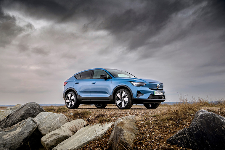 Side view of a blue Volvo electric vehicle parked on a gravel path, with a rocky foreground. The car is positioned against a dramatic sky with dark clouds, showcasing its modern design and sleek lines in a natural landscape.