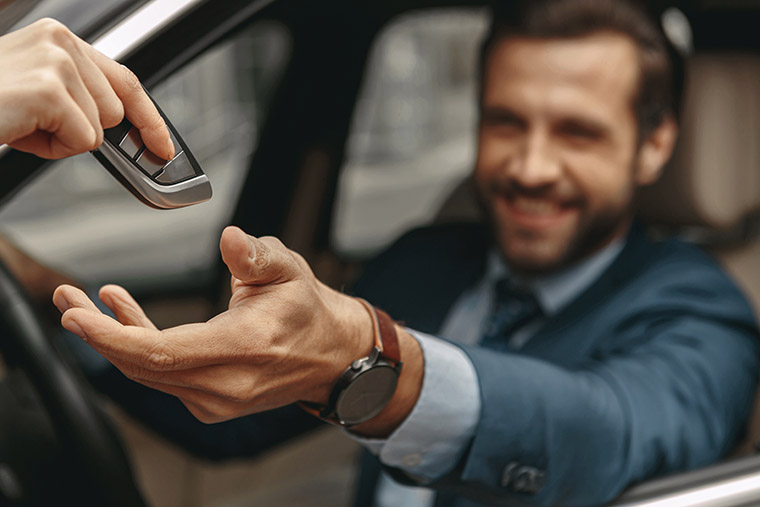 A close-up of a hand passing a car key to a smiling man in a blue suit, who is sitting in the driver's seat of a car. The man extends his hand to receive the key, and the image focuses on their hands with a blurred background.