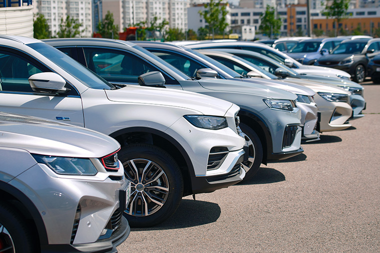 A row of silver and grey cars parked in a car dealership lot. The vehicles, mostly SUVs, are lined up in close proximity, showcasing their front ends with a clear blue sky and modern buildings in the background.