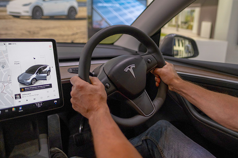 Close-up of hands gripping the steering wheel of a Tesla car, with the vehicle's central touchscreen showing navigation and music controls.