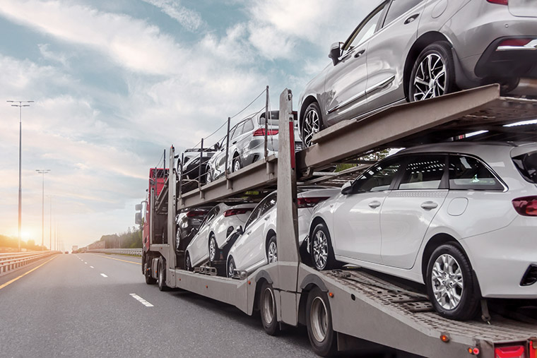 A car transporter truck driving on a highway, carrying multiple white vehicles stacked in two levels. The sky is partly cloudy, and the road stretches into the distance with lampposts visible along the side.