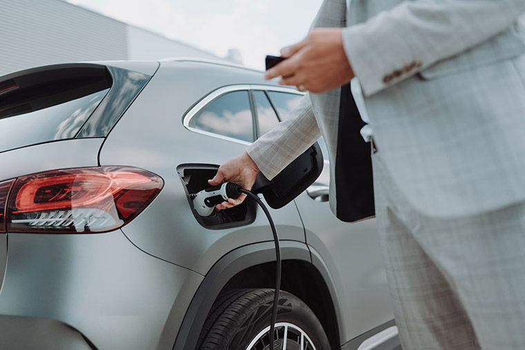 A man in a light grey suit holding an electric car charging cable, plugging it into the charging port of a silver electric vehicle. The car’s rear light is visible, and the background features a modern building and clear sky.