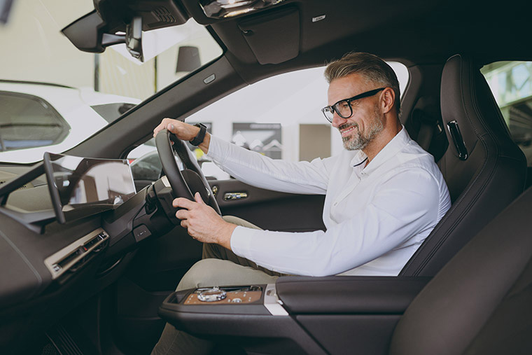 A middle-aged man with glasses smiling while seated in the driver's seat of a car. He is wearing a white shirt and light pants, with a large digital display visible on the dashboard. The car interior is modern and clean, with a bright showroom background.